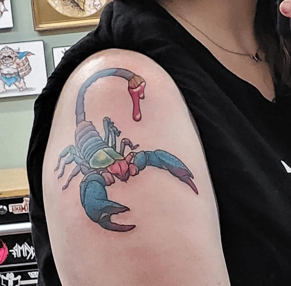 A rainbow-colored scorpion tattoo on a woman's upper arm, close to the shoulder. The scorpion has a paintbrush instead of a stinger, with magenta paint dripping from it.